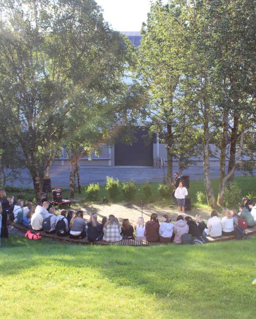 Bilde av elever som sitter i amfiet i bibliotekhagen på første skoledag 2022. Trær i bakgrunnen og Berit Aasberg som holder velkomsttale. - Klikk for stort bilde